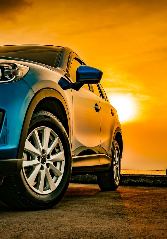 SUV Bleu Éclatant au Coucher de Soleil – Scène Automobile SUV bleu dynamique au coucher de soleil, lumière orange vibrante sur le véhicule et le ciel nuageux. Gros plan sur la jante et la route.